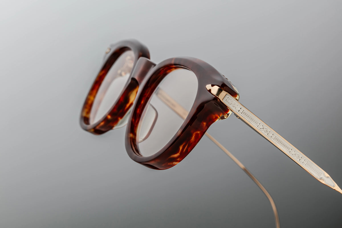 Macro close-up of the Jacques Marie Mage Hisao Breccia temple tips from Wink Optometrists. The shot details the lightweight gold beta titanium arms ending in custom burgundy epoxy tips with subtle, native-inspired hairline engravings.