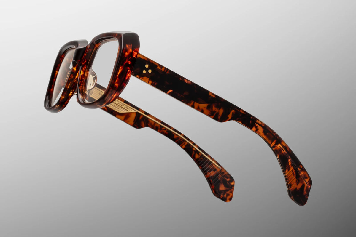 Macro close-up shot focusing on the temple arm of the Jacques Marie Mage Aldo RX in Argyle. The crystal-clear inner layer of the double-laminated acetate fully exposes a gold-toned, intricately engraved custom metal wire core. The detailed native-inspired engraving is visible alongside the robust, silver-polished 7-barrel hinge and the signature Burgundy domed 3D metal cameo icon embedded at the temple tip. Available at WINK Optometrists.
