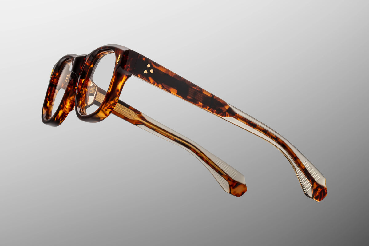 Side profile view of the Jacques Marie Mage Amilcar RX in Argyle. The thick, custom double-laminated acetate temple arms transition seamlessly from a deep Havana tortoiseshell exterior to a clear beige interior laminate. A custom 7-barrel hinge with refined, dark gold hairline details anchors the side arms firmly to the front frame. Available at WINK Optometrists.