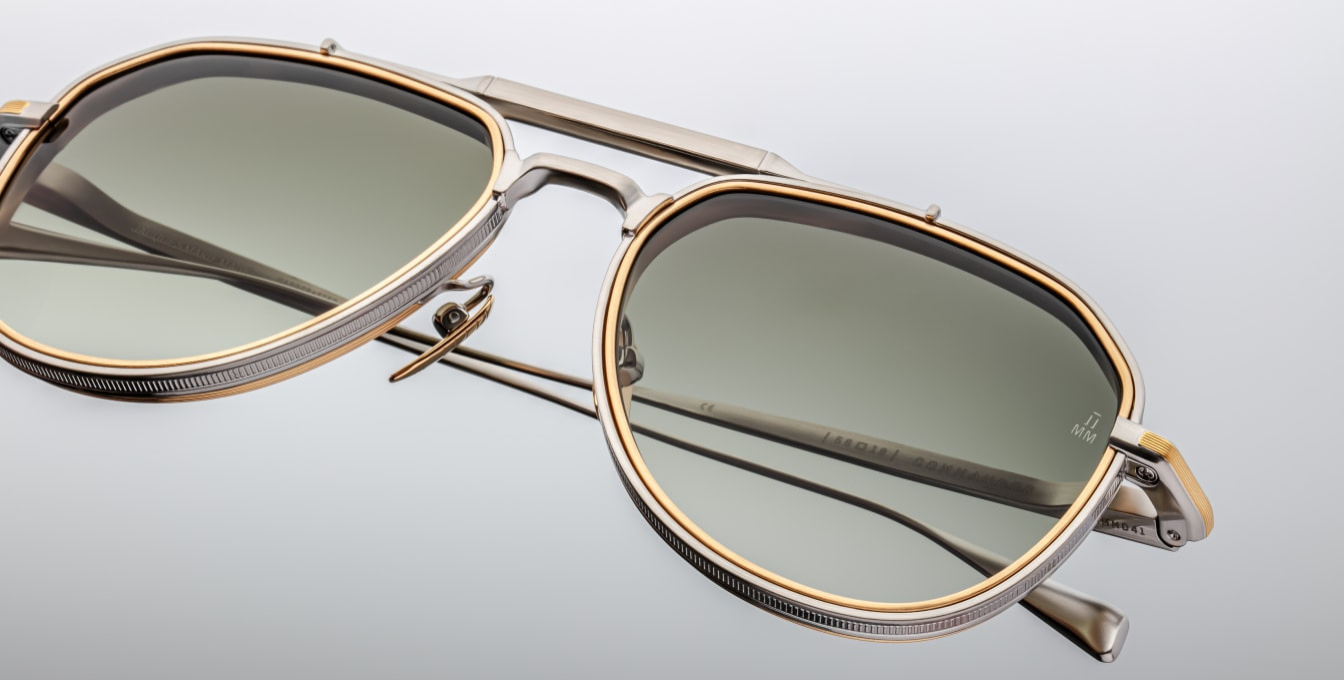 Close-up of the Jacques Marie Mage Commander Silver sunglasses, highlighting the 6mm titanium lens-rim inserts and dual-tone silver and dark gold beta titanium frame. Features include a double nose bridge, silver hairline-engraved temples with wing tips, custom tension-secured hidden hinges, and custom nose pads with engraved detailing. The light bottle green CR39 lenses offer 100% UV protection and backside anti-reflective treatment, combining vintage '70s aviator style with modern craftsmanship.