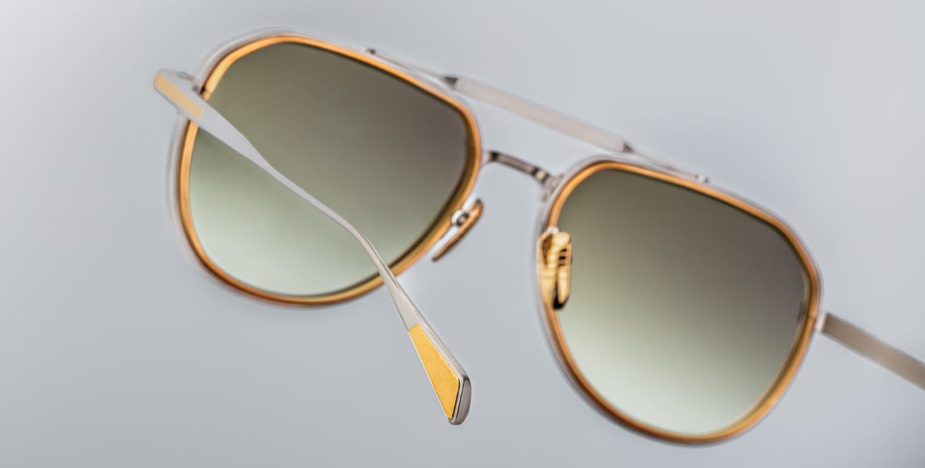 Close-up of the Jacques Marie Mage Commander Silver sunglasses, highlighting the 6mm titanium lens-rim inserts and dual-tone silver and dark gold beta titanium frame. Features include a double nose bridge, silver hairline-engraved temples with wing tips, custom tension-secured hidden hinges, and custom nose pads with engraved detailing. The light bottle green CR39 lenses offer 100% UV protection and backside anti-reflective treatment, combining vintage '70s aviator style with modern craftsmanship.