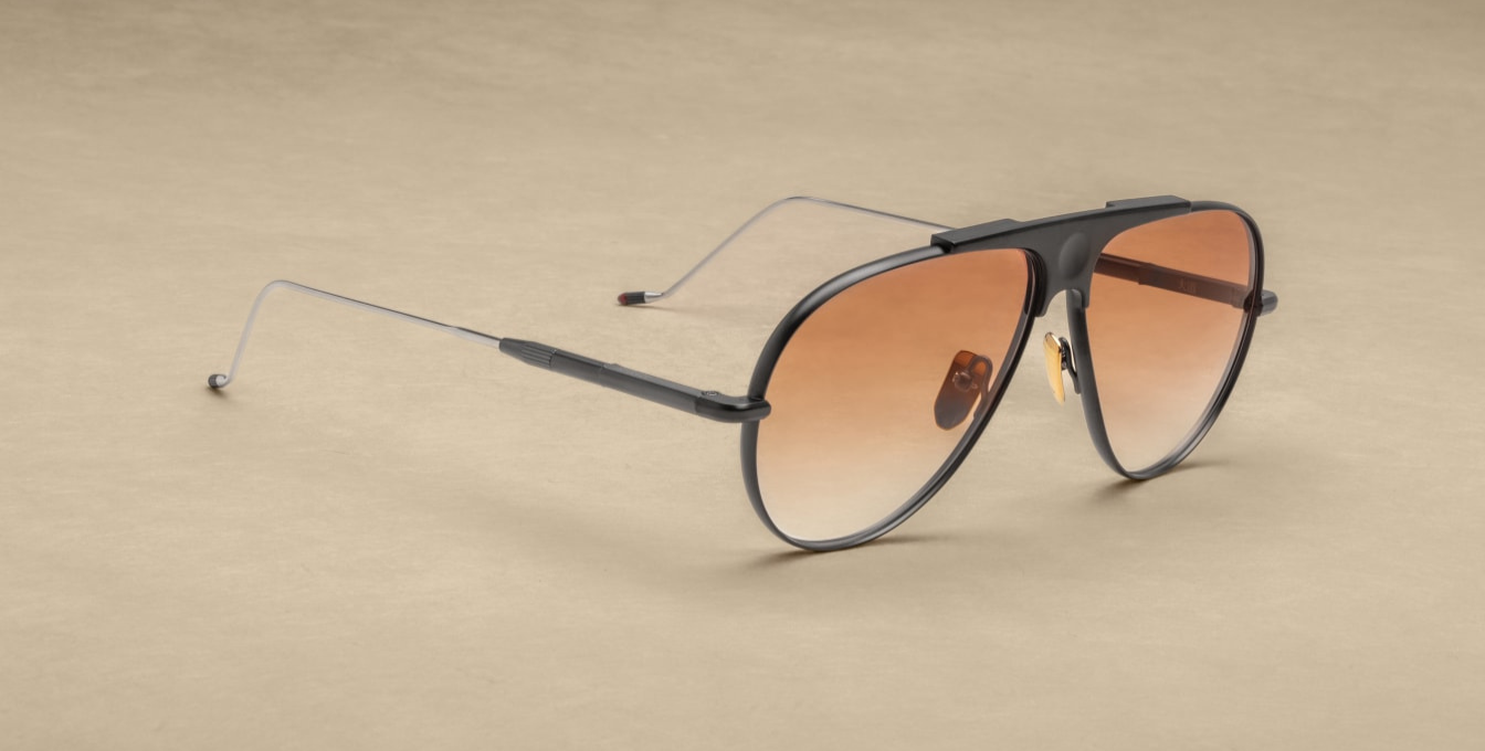 Jacques Marie Mage Daido sunglasses in Black, featuring a sculptural black and silver beta-titanium frame with a distinctive recessed bridge and circular depression. The design includes Art Deco-inspired hairline detailing, custom dark gold nose pads with engraved insignia, and tension-secured custom monoblock hinges. The apricot gradient CR39 lenses offer 100% UV protection and backside anti-reflective treatment, combining vintage aviator aesthetics with modern craftsmanship