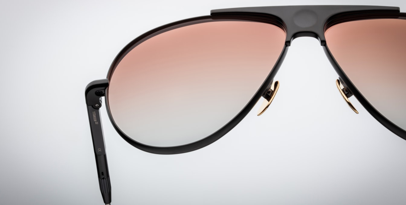 Jacques Marie Mage Daido sunglasses in Black, featuring extendable silver-tone beta titanium temples with burgundy epoxy tips and engraved detailing. The frame includes a distinctive recessed bridge with a circular depression and Art Deco-inspired hairline accents. Handcrafted in Japan, these limited-edition aviators offer 100% UV protection with apricot gradient CR39 lenses and backside anti-reflective treatment