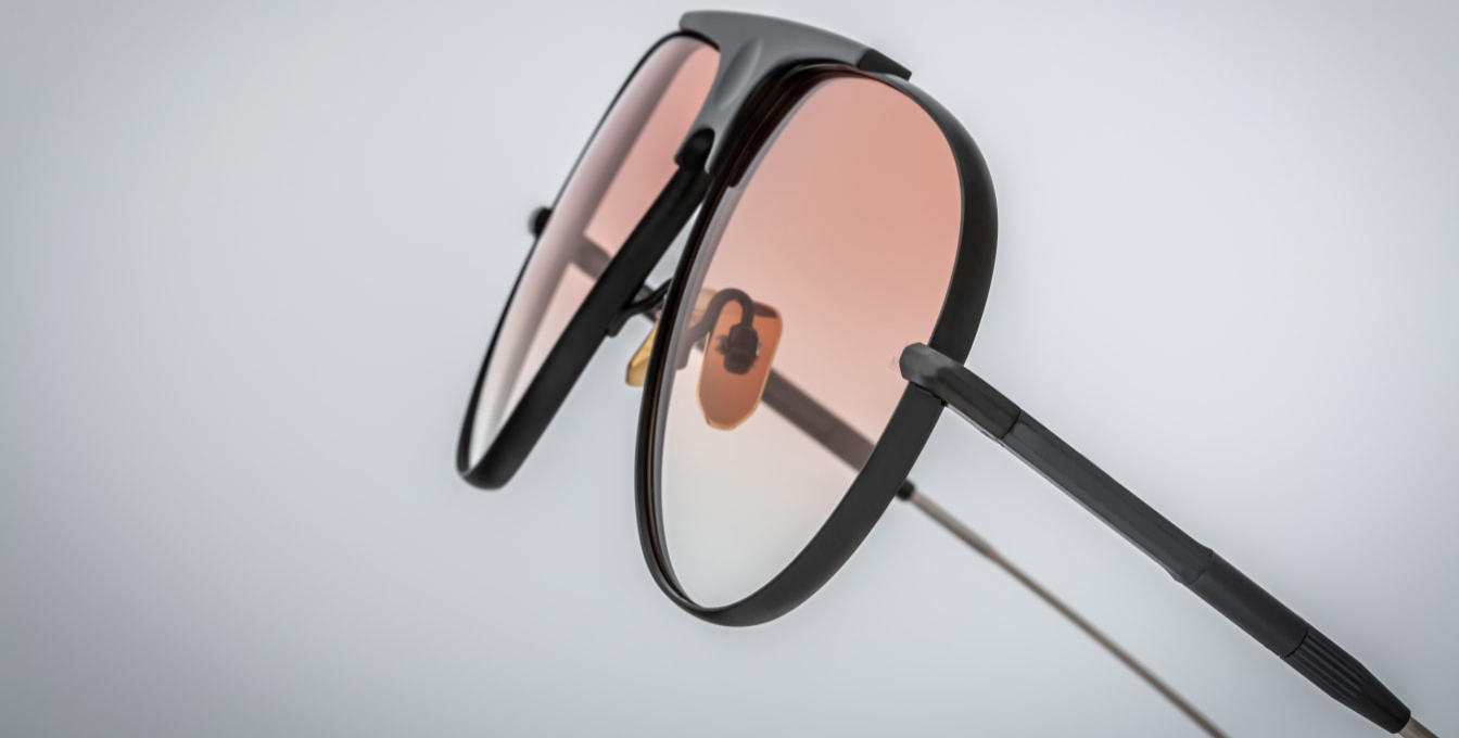 Close-up of the apricot gradient CR39 lenses in Jacques Marie Mage Daido Black sunglasses, offering 100% UV protection and backside anti-reflective treatment. The lenses are set within a sculptural black and silver beta-titanium frame, featuring a distinctive recessed bridge with a circular depression, Art Deco-inspired hairline detailing, and custom dark gold nose pads with engraved insignia.