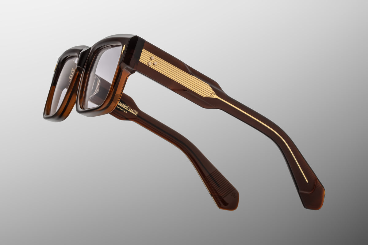 Close-up detail of the temple arm on Jacques Marie Mage Hemmings Café RX optical glasses. The crystal-clear outer laminate layer reveals an intricate, custom hairline-engraved gold wire core running through the deep brown interior arm.