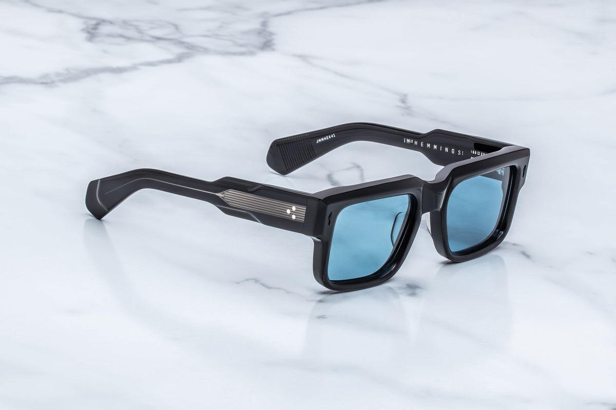 A side-profile studio photograph focusing on the temple arm of the Jacques Marie Mage Hemmings sunglasses in the Mystique colourway. The chunky 10mm dual-laminated acetate construction transitions from a matte black exterior on the front frame to a crystal-clear, polished transparent acetate layer along the temple. This transparent layer completely exposes a highly detailed, sterling silver palladium core wire featuring intricate, Art Deco-inspired hairline engravings. A multi-barrel custom hinge securely f