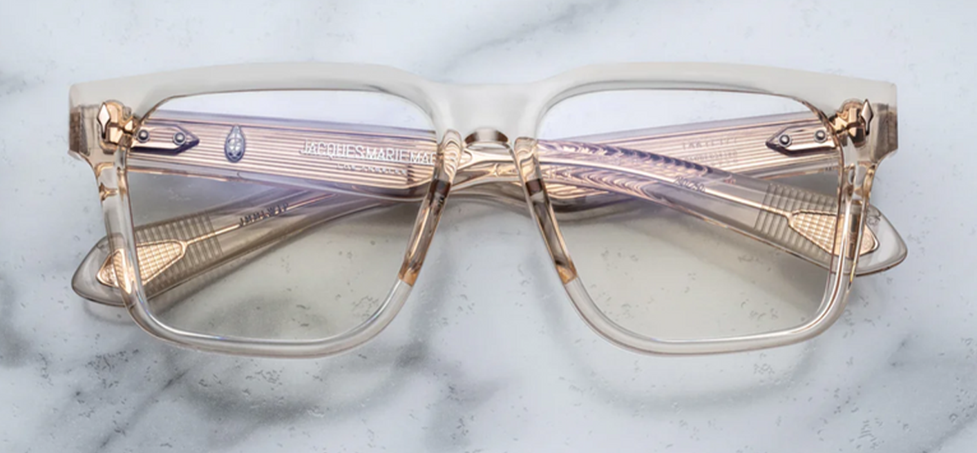 front view of Jacques Marie Mage Lucknow Pearl glasses in translucent crystal-acetate . The bold rectangular frame is 10 mm thick with sculpted volume cuts and a flat browline. At each hinge front, a polished light-gold sterling arrowhead pin adds Art Deco flair. The lenses are super-light bronze-tinted CR39 with backside anti-reflective coating and full UV protection.