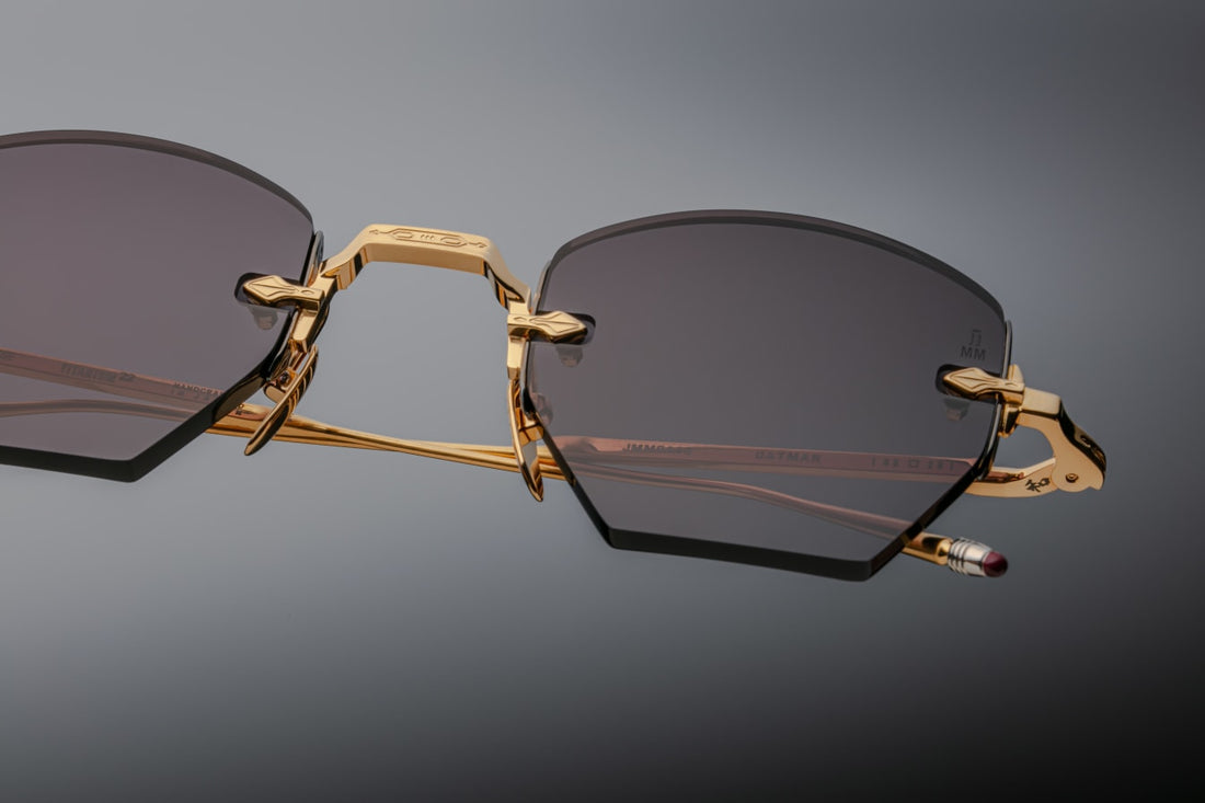 Macro view of the cool-toned charcoal-grey lens nestled into the gold frame: hexagonal lens edge with crisp polished bevel, and arrowhead filigree affixed at the rim’s midpoint. Visible through the lens is the JMM logo subtly etched onto the nose pad arm.