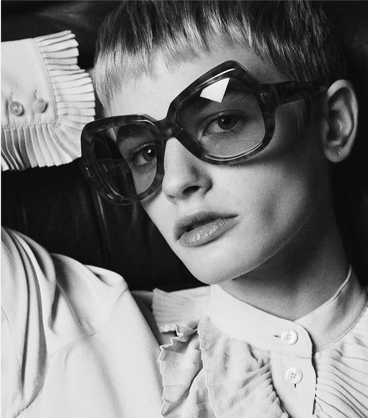 Model wearing Jacques Marie Mage Perreti Camel: oversized square‑round acetate frame in warm honey‑brown tortoiseshell, sitting slightly oversized on the face. The full‑rim front features a sculpted exaggerated brow line, a softly rounded saddle bridge, and two light‑gold arrowhead pins at the upper outer corners. Mint‑green gradient CR‑39 lenses are visible through the frame, providing a soft contrast to the camel tones