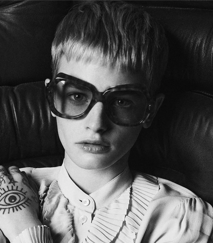 Model wearing Jacques Marie Mage Perreti Camel: oversized square‑round acetate frame in warm honey‑brown tortoiseshell, sitting slightly oversized on the face. The full‑rim front features a sculpted exaggerated brow line, a softly rounded saddle bridge, and two light‑gold arrowhead pins at the upper outer corners. Mint‑green gradient CR‑39 lenses are visible through the frame, providing a soft contrast to the camel tones