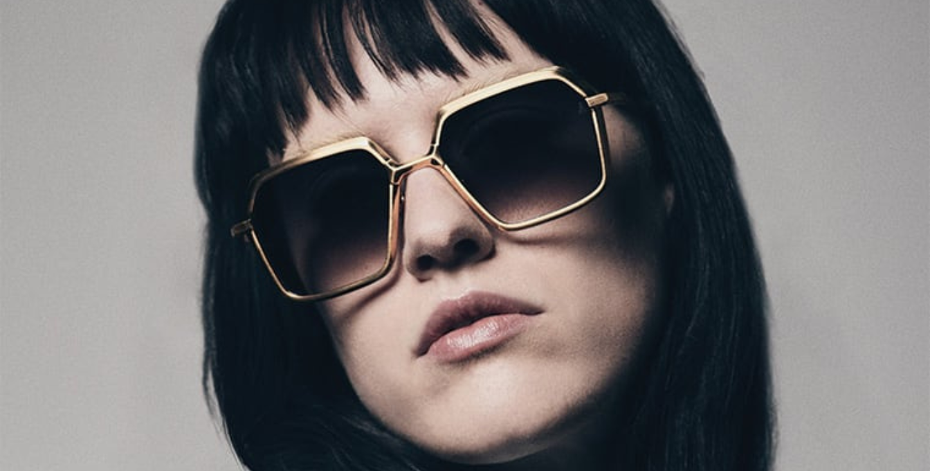model wearing Jacques Marie Mage Ugo Gold sunglasses, showcasing the gold-toned beta titanium frame with sculpted hexagonal volumes and hairline epoxy detailing. The acetate temple tips with exposed wirecore details are prominently displayed