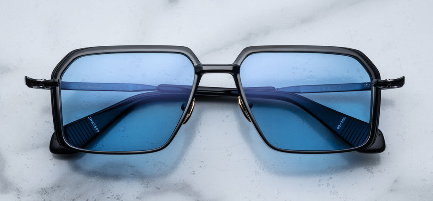 Front view of Jacques Marie Mage Vasco Black sunglasses, featuring a sharp rectangular silhouette with a V-shaped profile. The frame is crafted from black titanium, showcasing sculpted volume cuts and hairline epoxy detailing. Acetate temple tips with exposed wirecore details complement the design. The azul CR39 lenses offer 100% UV protection and backside anti-reflective treatment