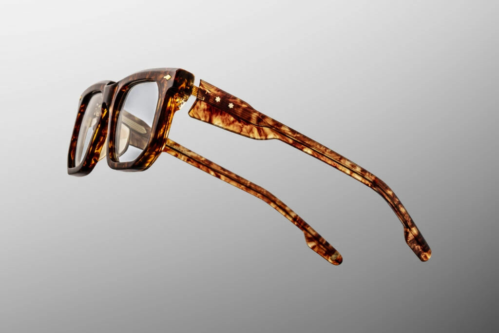 Side profile view of the Jacques Marie Mage eyeglasses in Argyle. The focus is on the thick temple arms which transition from a rich Havana tortoiseshell exterior to a clear beige interior laminate. A premium, custom dark gold tension-secured multi-barrel hinge firmly fastens the temple arm to the front frame face. Available at WINK Optometrists.
