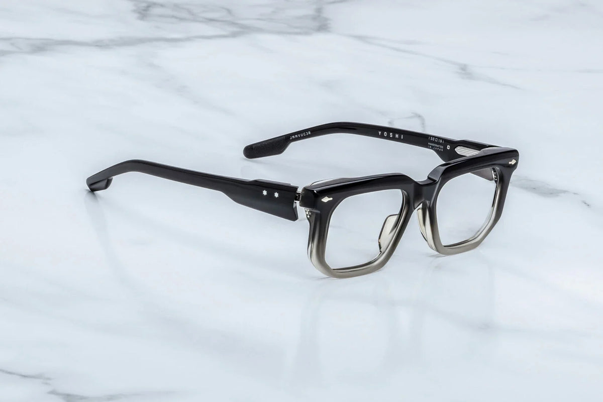 Three-quarter studio shot of the Jacques Marie Mage Yoshi Black Fade RX glasses. The perspective highlights the heavy profile depth of the geometric frame face and its hand-polished architectural edges. The striking black-to-clear gradient effect of the rims is visible under the studio lighting, flowing smoothly into custom double-laminated temples. Available at WINK Optometrists.