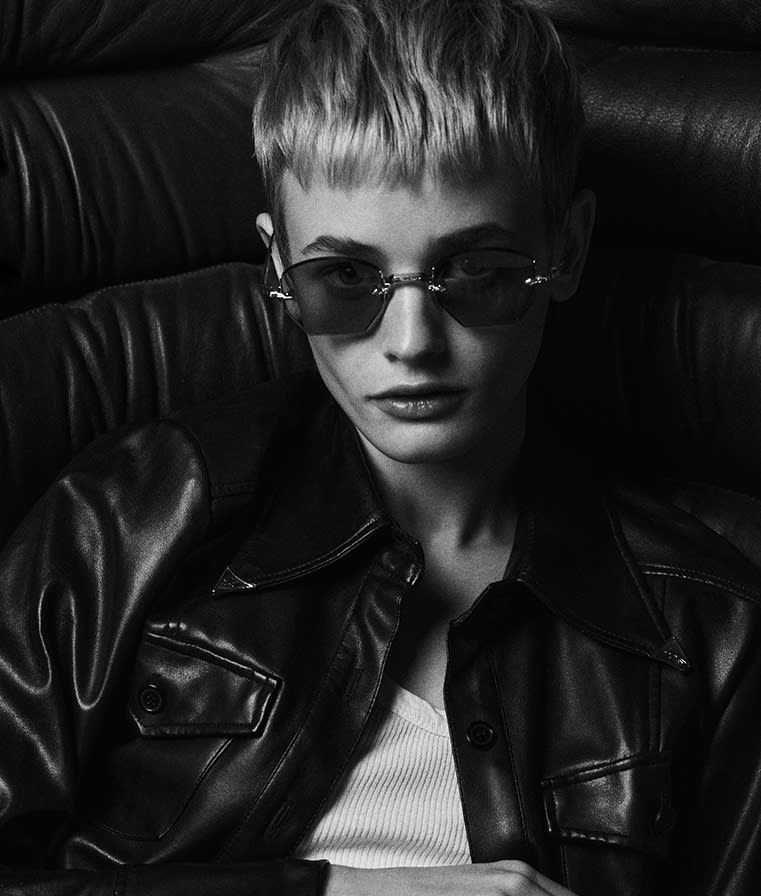A gender-neutral model wearing Jacques Marie Mage Oatman Gold: the rimless gold hexagonal lenses sit perched above the nose, with delicate gold temples extending back. The unique tailfin tips peek past the ears. The sunglasses cast a refined yet boldly minimalist statement against the model’s neutral outfit.