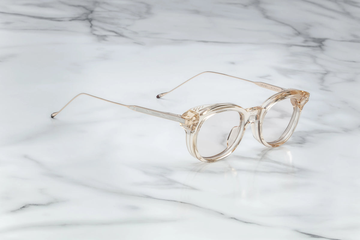  Profile view of the Jacques Marie Mage Hisao Pearl frame. The translucent clear acetate front leads to contrasting 100% beta titanium temples engineered in a dual-toned light gold and silver finish. The sleek metal arms feature custom, native-inspired horizontal line engravings, terminating in custom-molded temple tips dipped in JMM’s iconic burgundy bordeaux epoxy.