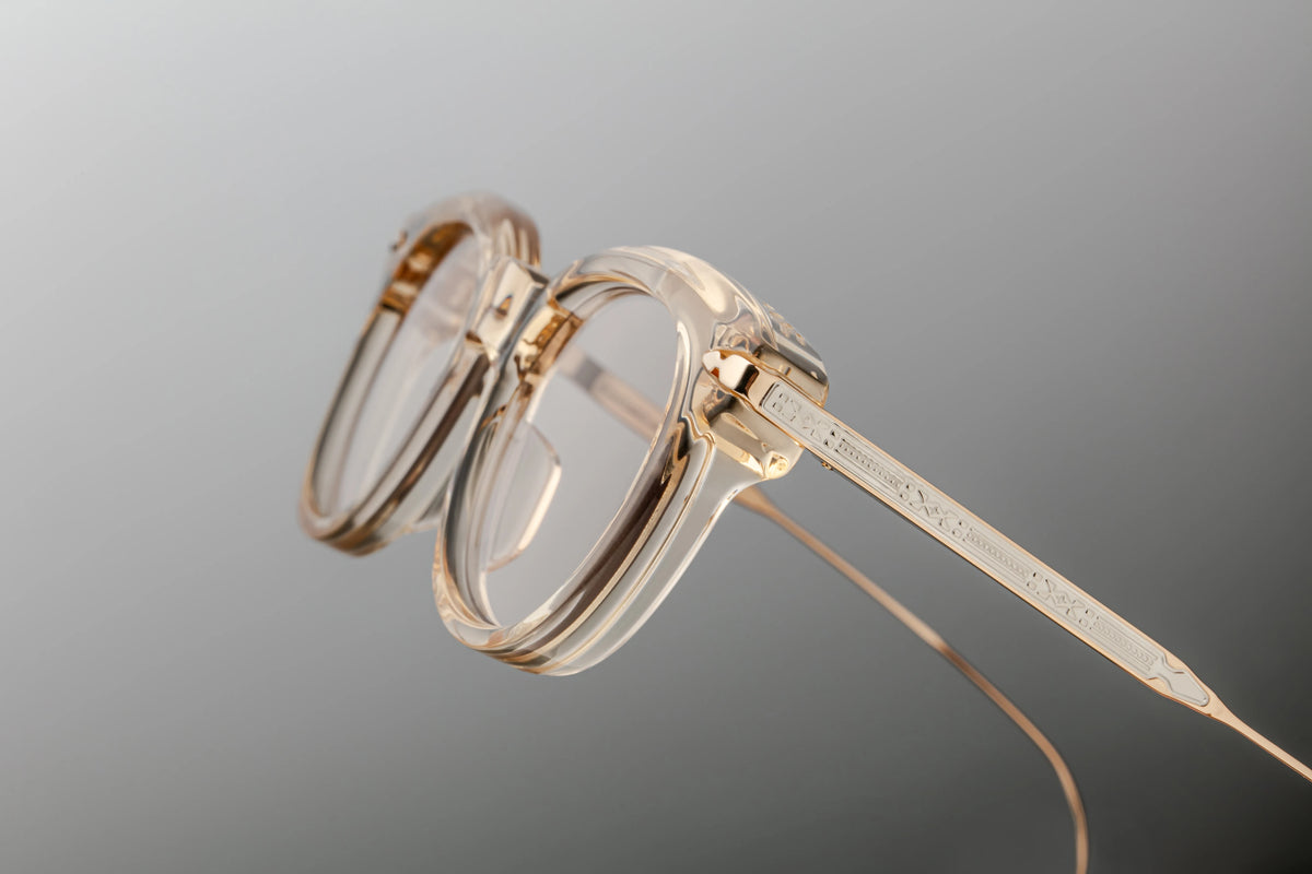 Three-quarter studio perspective of the Jacques Marie Mage Hisao Pearl optical frame. The angled shot emphasizes the crystalline depth and architectural bevelling of the thick, transparent front rim. The floating custom monoblock hinge system is completely visible through the acetate, showcasing the premium integration between the clear crystal front and the engraved light-gold titanium