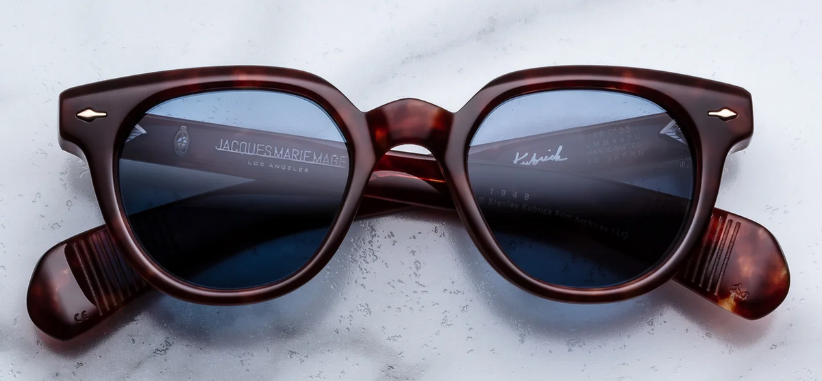 Front view of Jacques Marie Mage Stanley Kubrick 1948 sunglasses in Breccia. Features a bold, thick, rounded mid-century acetate frame with a striking marbled dark brown and Havana texture. Includes a structured keyhole nose bridge, signature sterling silver arrowhead front pins, and rich Prussian blue lenses