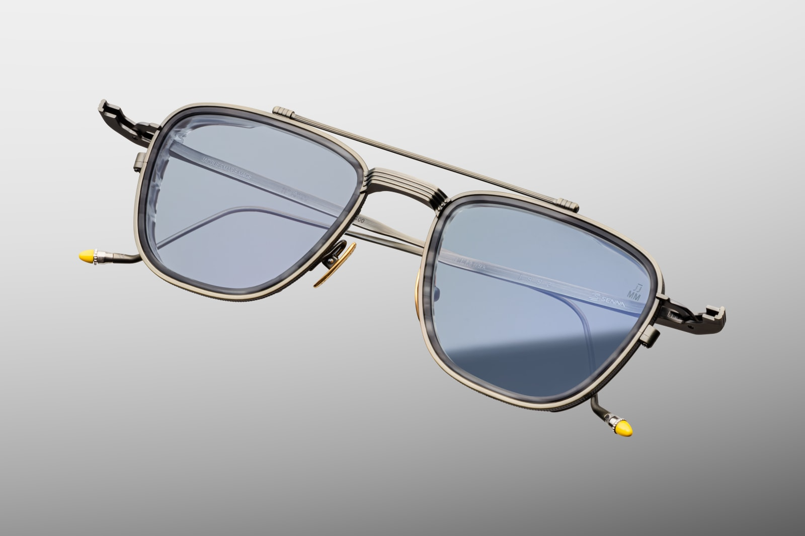 Close-up of front of Jacques Marie Mage Ayrton Senna 1992 Antique Gunmetal sunglasses highlighting the sculptural top-bar, coined-edge eyerims, polished gold nose-pads and light blue crown-mineral lenses, available at WINK Optometrists.