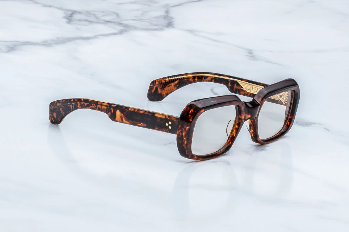 Three-quarter studio shot of the Jacques Marie Mage Aldo RX Argyle glasses. The perspective highlights the substantial depth of the 10mm thick cured acetate frame and the crisp, beveled edges along the top brow. The clear demo lenses reveal the chunky profile. The custom double-laminated temple is visible, showcasing the transition from the rich tortoise front to the side construction. Available at WINK Optometrists.