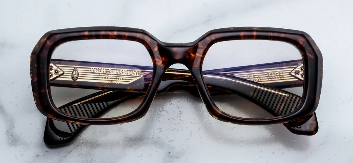 Front view of Jacques Marie Mage Aldo RX eyeglasses in Argyle. The frame features a bold, sculptural, oversized 1970s-inspired rectangular silhouette with deep lenses and a distinctively beveled brow. The 10mm block acetate showcases a rich tortoise pattern blending dark brown, amber, and translucent spots. Signature silver arrowhead front pins accent the top outer corners of the frame.
WINK Optometrists