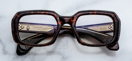 Front view of Jacques Marie Mage Aldo RX eyeglasses in Argyle. The frame features a bold, sculptural, oversized 1970s-inspired rectangular silhouette with deep lenses and a distinctively beveled brow. The 10mm block acetate showcases a rich tortoise pattern blending dark brown, amber, and translucent spots. Signature silver arrowhead front pins accent the top outer corners of the frame.
WINK Optometrists