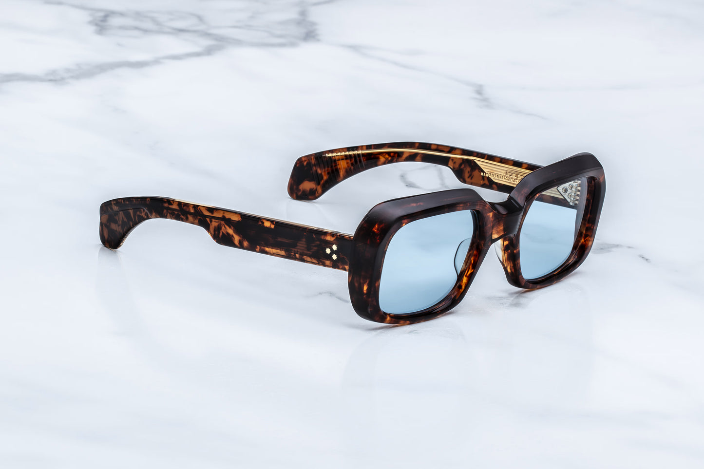  Three-quarter studio shot of the JMM Aldo Argyle sunglasses, highlighting the distinctively beveled brow line, thick 10mm acetate build, and striking blue lens tint