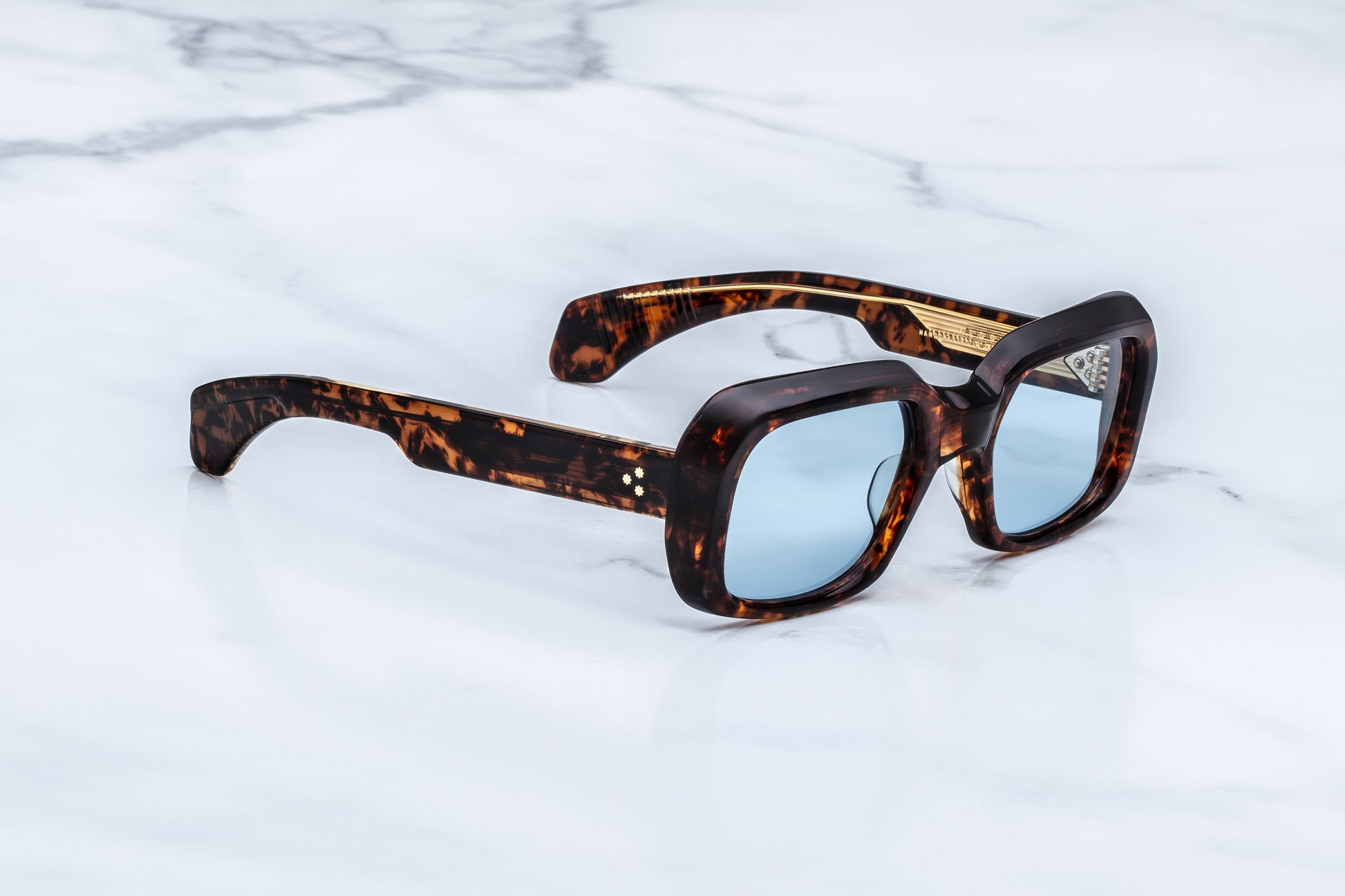  Three-quarter studio shot of the JMM Aldo Argyle sunglasses, highlighting the distinctively beveled brow line, thick 10mm acetate build, and striking blue lens tint