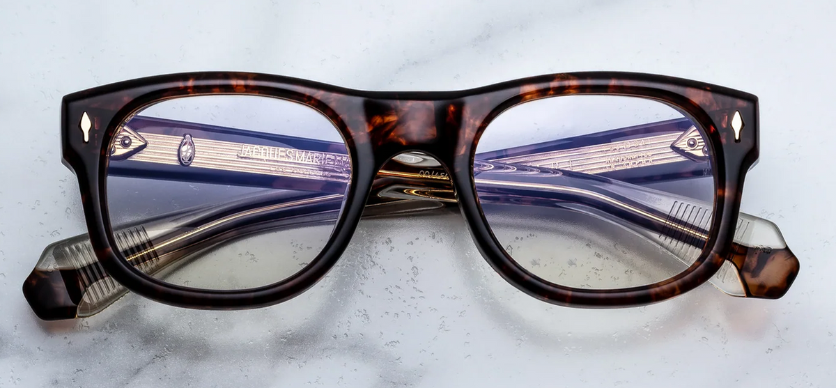  Front view of Jacques Marie Mage Amilcar RX eyeglasses in Argyle. The frame features a classic round lens shape paired with a softly arched, grand touring-inspired brow line. Handcrafted from substantial 10mm cured cellulose acetate in a rich Havana tortoiseshell pattern, it is elevated by 18K dark gold signature arrowhead front pins at the outer top corners. Available at WINK Optometrists.