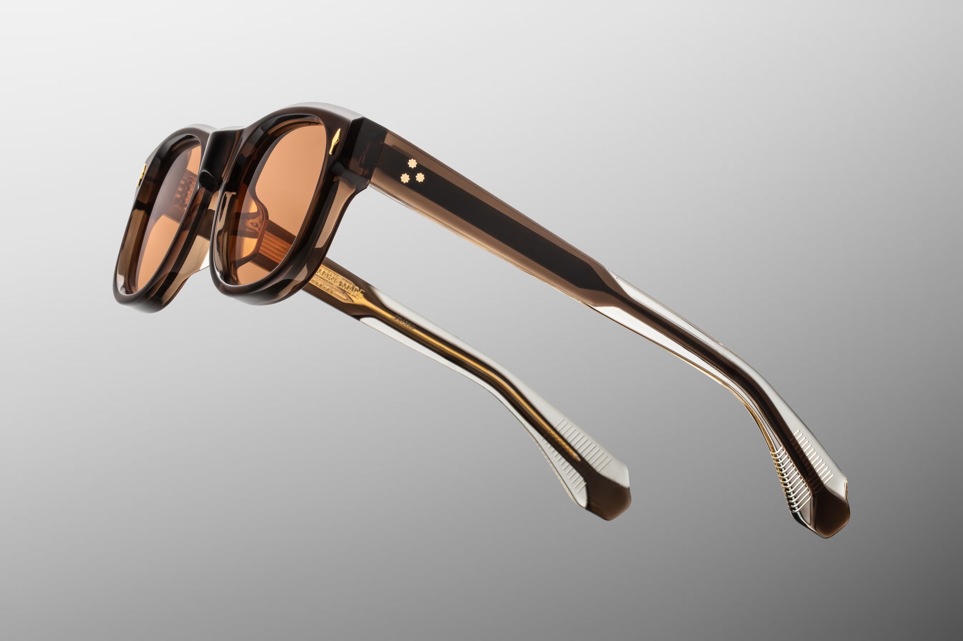 Side view of the JMM Amilcar sunglasses temple arm, showcasing the polished brown exterior finish, 10k gold spur-shaped rivet heads, and the dark gold wirecore running through the clear beige inner laminate.