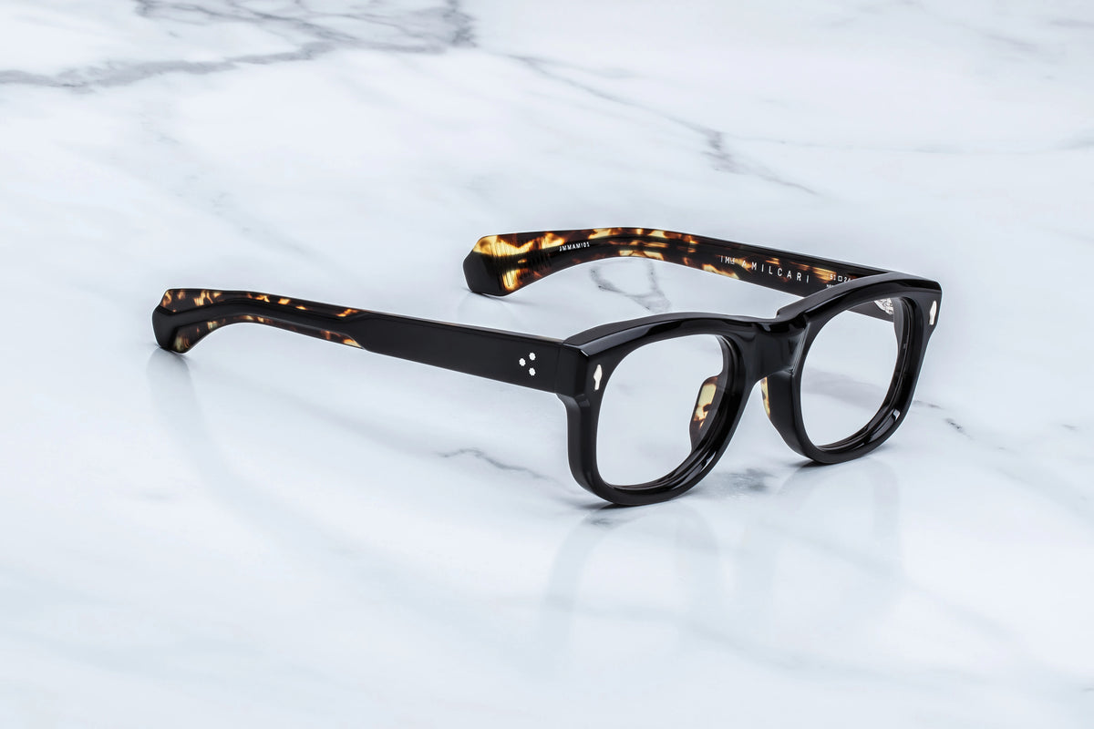  Three-quarter studio shot of the Jacques Marie Mage Amilcar RX Noir eyeglasses. The perspective showcases the heavy 10mm profile depth of the glossy black frame face alongside its smooth, sculpted contours. The clear demo lenses highlight the structural thickness of the rims, transitioning seamlessly into the double-laminated temples. Available at WINK Optometrists