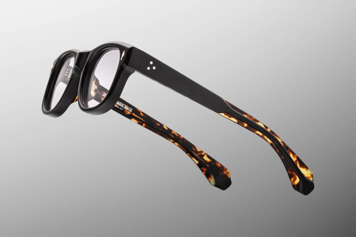 Macro close-up shot focusing on the inside temple arm of the Jacques Marie Mage Amilcar RX in Noir. The polished translucent Havana tortoiseshell inner layer fully exposes the custom 'Viceroy' metal wire core. The metal core is intricately laser-engraved with geometric, refined Art Deco hairline patterns. Gold foil-stamped hallmarks are cleanly pressed into the acetate near the secure 7-barrel hinge mechanism. Available at WINK Optometrists