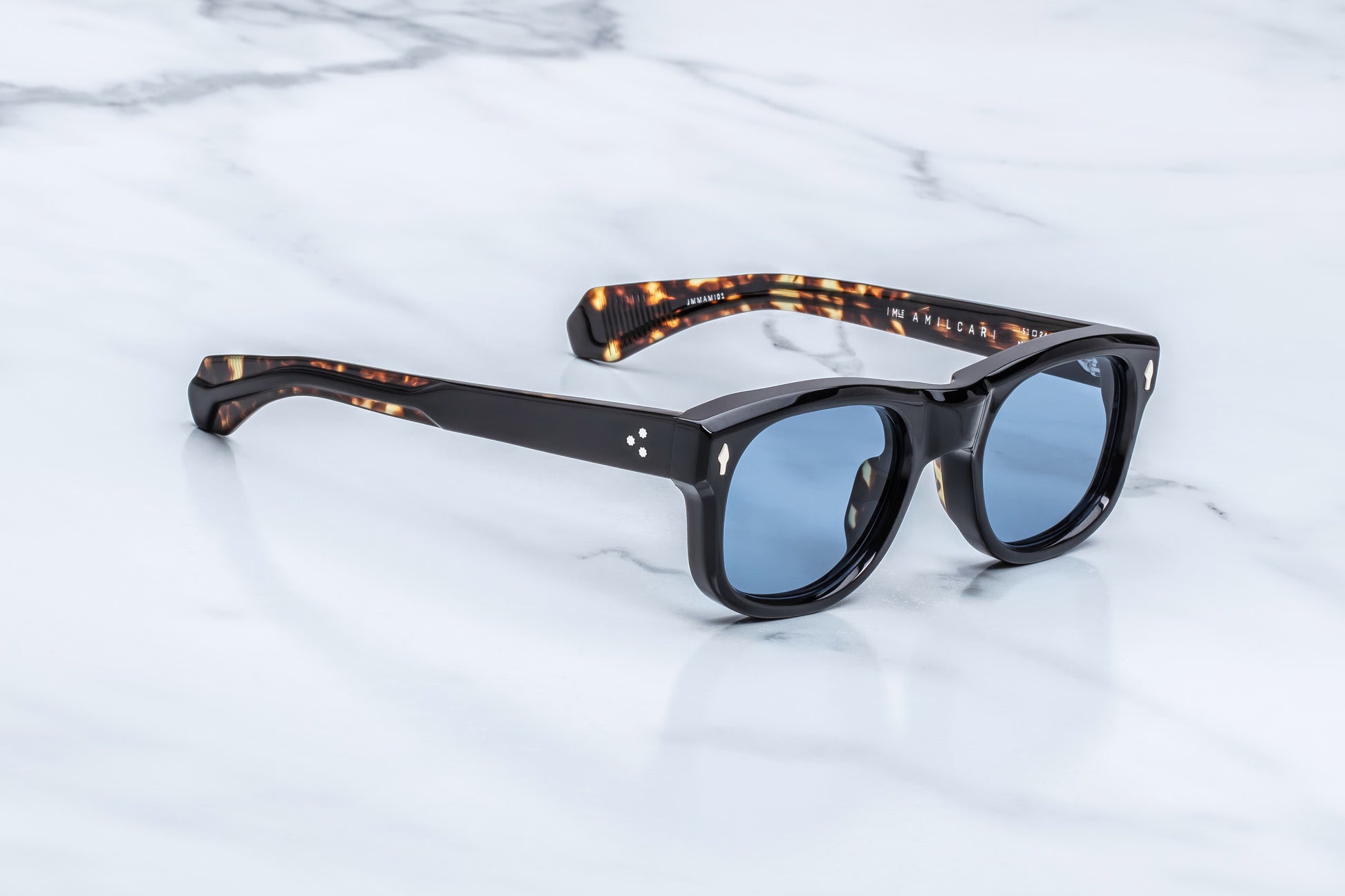 Three-quarter studio shot of the JMM Amilcar Noir sunglasses, highlighting the substantial 10mm thick block frame, deeply recessed cerulean blue lenses, and sculpted vintage-inspired silhouette