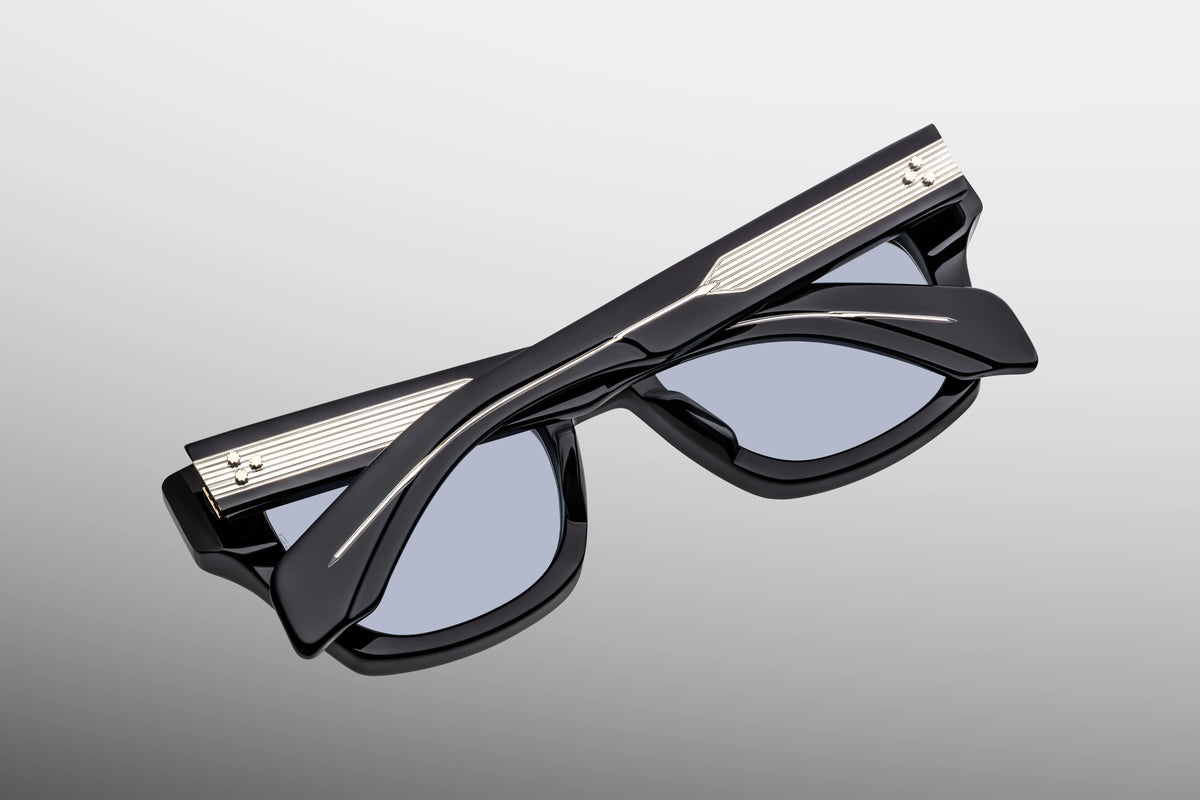 Detailed close-up view from behind the hinge of the JMM Dealan 53 Obscure sunglasses. The perspective reveals a heavy-duty, tension-secured custom 7-barrel hinge in a dark gold finish. On the inner temple tip, a dark gold domed 3D metal cameo logo is visible against the smooth, deep blue acetate