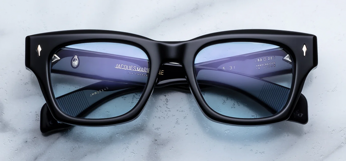 A front-facing product shot of Jacques Marie Mage Dealan 53 sunglasses in the Obscure colourway. The oversized, 10mm structured rectangular acetate frame with a subtle cat-eye flare is crafted from a rich, translucent deep ink-blue. Serene blue lenses are housed inside the substantial frame, flanked by iconic sterling silver arrowhead pins embedded horizontally on the upper outer corners.