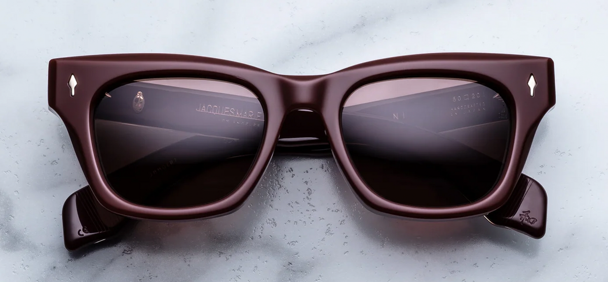  Front view of Jacques Marie Mage Dealan sunglasses in Bordeaux Empire. The frame displays a bold, 1960s-inspired rectangular silhouette featuring a subtle cat-eye lift and strong, confident proportions. The 10mm block acetate is a deep, polished cassis burgundy-red, housing striking ruby-tinted lenses. Signature sterling silver arrowhead front pins with custom hand-painted bordeaux epoxy enamel inlays accent the upper outer corners. Available at WINK Optometrists