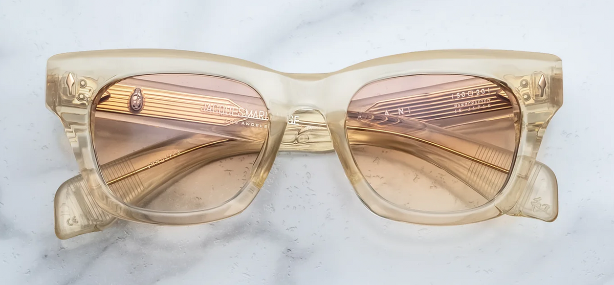 Front view of Jacques Marie Mage Dealan sunglasses from the Photochromic Collection in Halo. The frame displays a bold, 1960s-inspired rectangular silhouette featuring a subtle cat-eye lift and strong, chunky volumes. The 10mm block acetate is a translucent Polished Brut beige, housing responsive photochromic brown gradient lenses. Signature Light Gold arrowhead front pins accent the upper outer corners. Available at WINK Optometrists