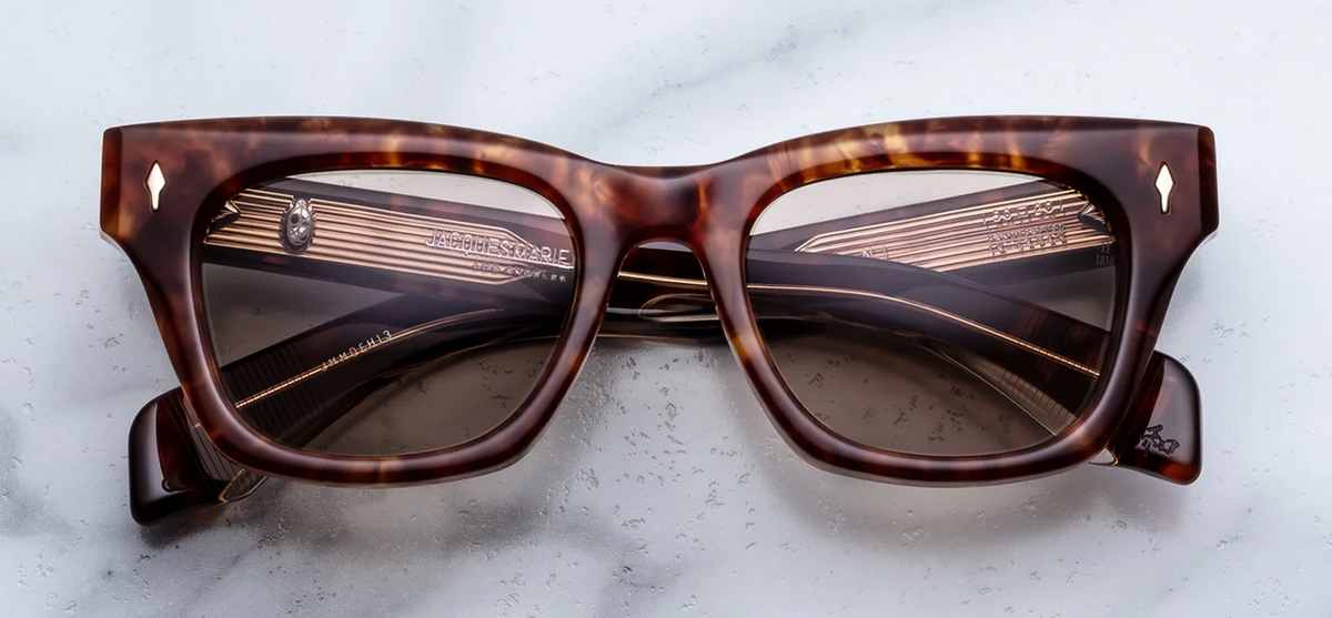 A front-facing product shot of Jacques Marie Mage Dealan sunglasses in Honeycomb, size 50/20. The medium-fit, 10mm block cellulose acetate frame features structured rectangular lines with a subtle cat-eye angle in a warm, translucent amber-honey and dark brown Havana tortoiseshell. Light gold arrowhead pins flank the solid brown lenses on the upper outer corners.