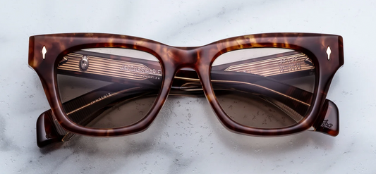 A front-facing product shot of Jacques Marie Mage Dealan sunglasses in Honeycomb, size 50/20. The medium-fit, 10mm block cellulose acetate frame features structured rectangular lines with a subtle cat-eye angle in a warm, translucent amber-honey and dark brown Havana tortoiseshell. Light gold arrowhead pins flank the solid brown lenses on the upper outer corners.