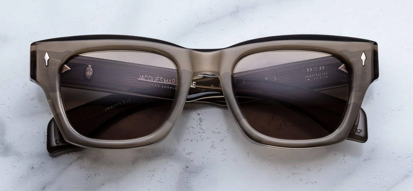 Front view of Jacques Marie Mage sunglasses in the Lescot colourway, featuring a dark charcoal-grey 10mm acetate frame, brass-toned lenses, and silver signature arrowhead front pins.