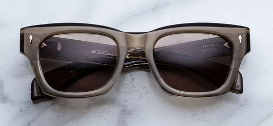 Front view of Jacques Marie Mage sunglasses in the Lescot colourway, featuring a dark charcoal-grey 10mm acetate frame, brass-toned lenses, and silver signature arrowhead front pins.