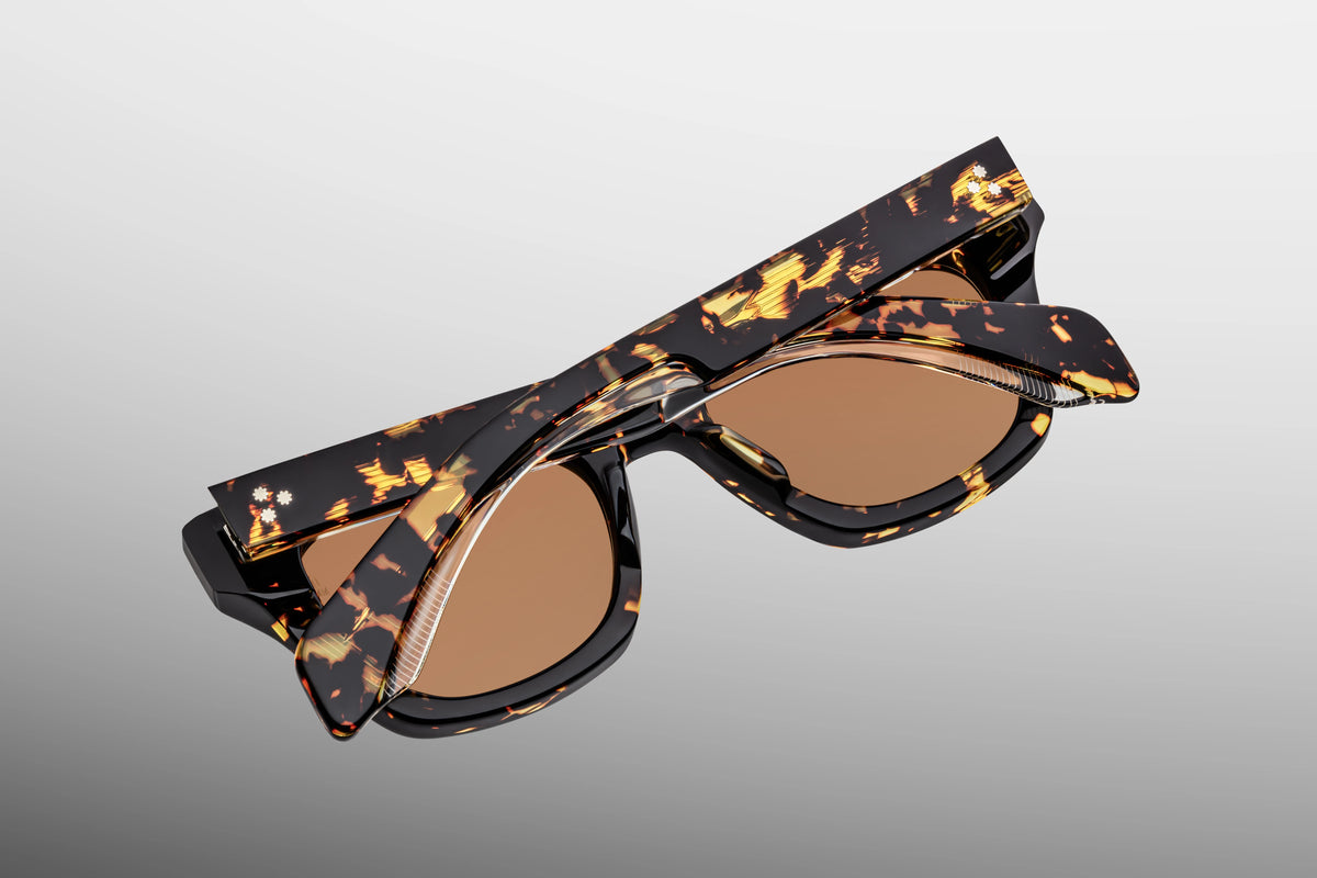 Detailed close-up view from behind the hinge of the JMM Dealan Tokyo sunglasses. The shot shows the internal "50 x 20" sizing engraving on the inner temple alongside a robust, custom 7-barrel tension-secured hinge in gold. A tiny, domed 3D metal cameo logo is visible near the ear piece tip against the dark patterned acetate