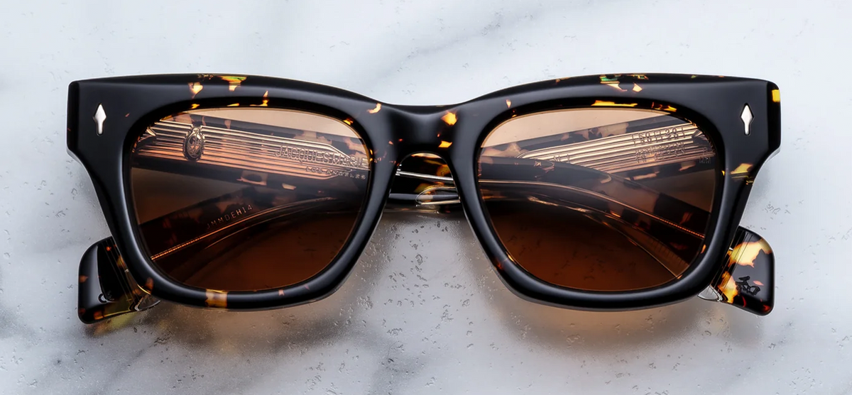 A front-facing product shot of Jacques Marie Mage Dealan 50/20 sunglasses in the Tokyo colourway. The medium-fit, 10mm structured rectangular acetate frame features a bold, classic dark Havana tortoiseshell pattern. Vibrant, solid orange lenses are framed by the dark mottled acetate, accented by signature dark gold or light gold arrowhead pins on the upper outer corners
