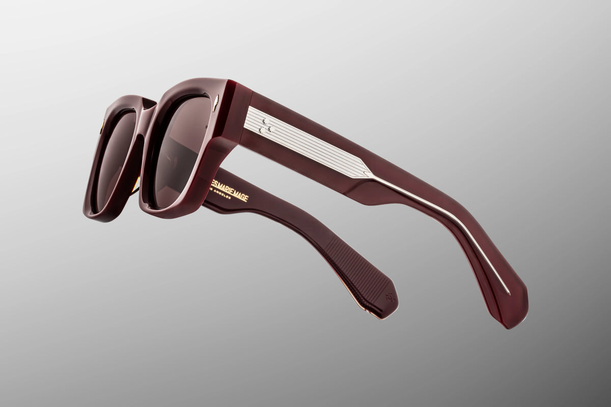 Macro shot of the temple arm on Jacques Marie Mage Enzo Bordeaux Empire sunglasses, emphasizing the interplay of light across the deep burgundy acetate surface and the exposed silver wire core with hairline filigree.