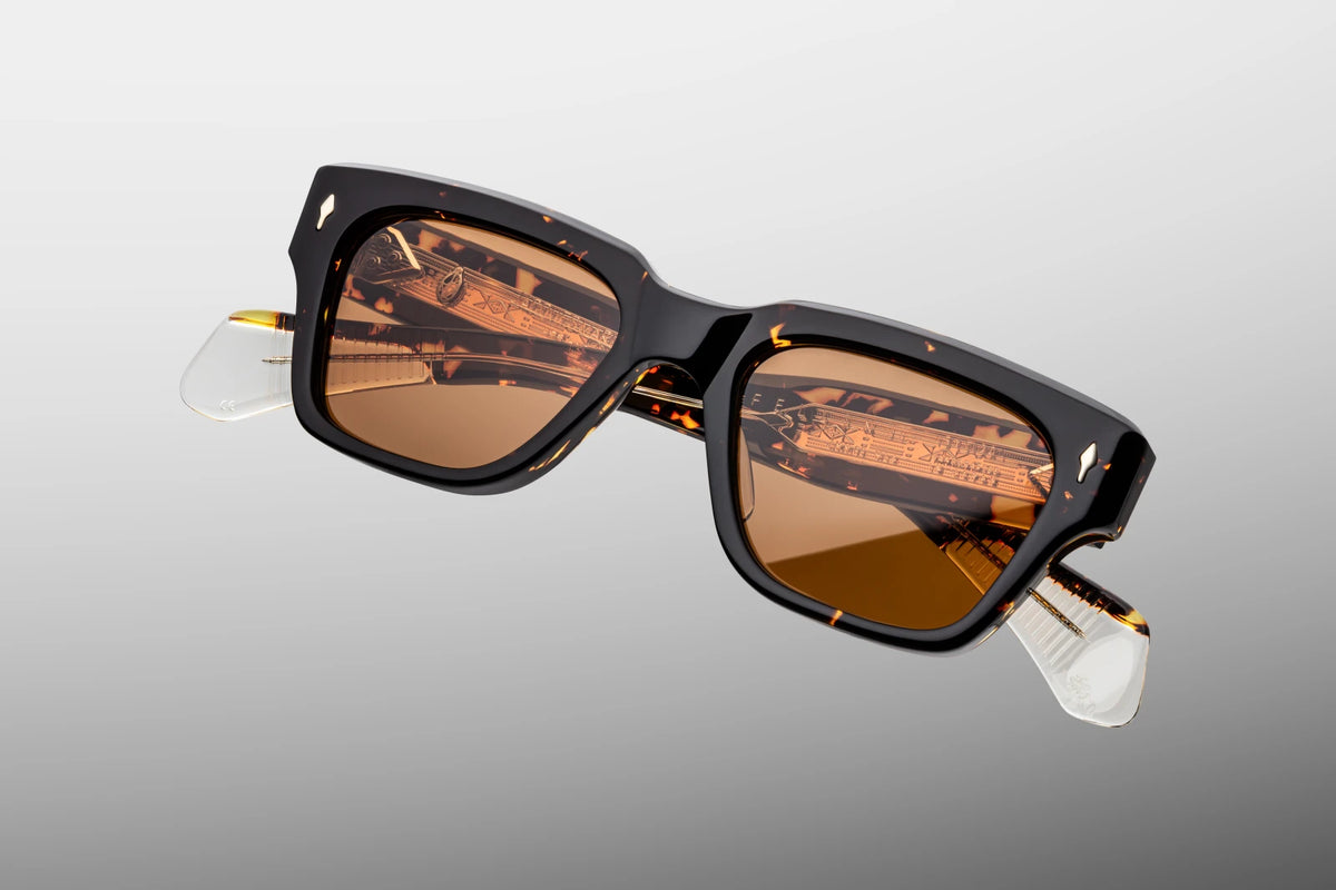A macro close-up focusing on the front upper corner of the JMM Fellini Tokyo frame. The shot details the hefty 10mm thick bevelled edge of the hand-polished tortoiseshell acetate. A sterling silver arrowhead front pin is embedded seamlessly flush into the frame, contrasting cleanly against the dark patterned acetate and the edge of the bright orange lens.
