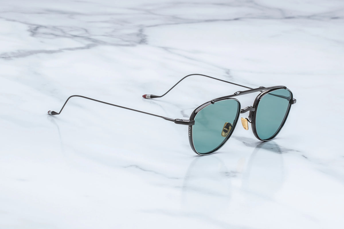 Three-quarter view of the Jacques Marie Mage Flynn Paon sunglasses. The angle highlights the sleek, dual-tone metal architecture of the black and silver titanium chassis and the thin wire temples bridging into the geometric frame front.