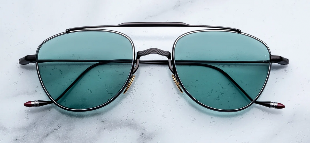 Front view of Jacques Marie Mage Flynn Paon sunglasses. Displays a compact, lightweight 100% beta titanium pilot aviator frame with a polished black double bridge, contrasting silver palladium eye rims, and vibrant turquoise solid-tint lenses.