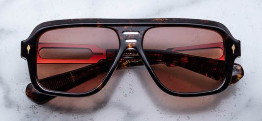 Jacques Marie Mage Fugue sunglasses in Agar, featuring a bold '60s-inspired navigator silhouette. The frame is crafted from 10mm cured cellulose acetate in a rich hickory hue, accented with silver arrowhead front pins and a distinctive double-bridge design. The amber CR39 lenses offer 100% UV protection and backside anti-reflective treatment