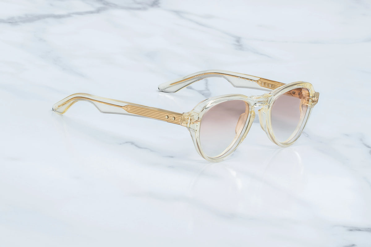 Three-quarter studio shot of the Jacques Marie Mage Hatfield Halo photochromic sunglasses. The perspective highlights the deep 10mm profile of the translucent champagne acetate rims and the softly curved contours of the aviator front. The photochromic brown gradient lenses show a subtle tint gradient, transitioning smoothly into matching clear-beige acetate temples. Available at WINK Optometrists.
