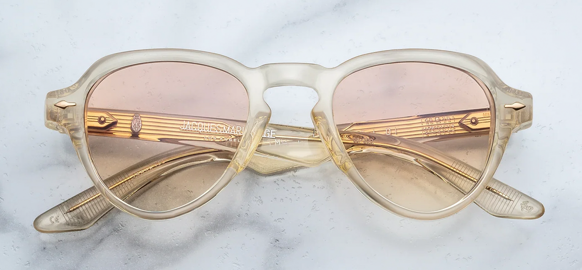 Front view of Jacques Marie Mage Hatfield sunglasses from the Photochromic Collection in Halo. The frames exhibit a compact, structured, retro-inspired aviator silhouette with a distinctively tailored lens shape. The heavy 10mm block acetate is a translucent Polished Brut champagne-beige, housing responsive photochromic brown gradient lenses. Signature Light Gold arrowhead front pins ornament the upper outer corners of the frame face. Available at WINK Optometrists.
